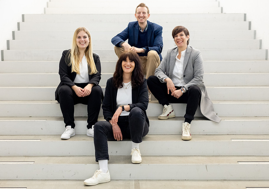 Vier Personen sitzen lächeln auf Stufen, alle tragen Business-Casual-Kleidung und Sneakers.